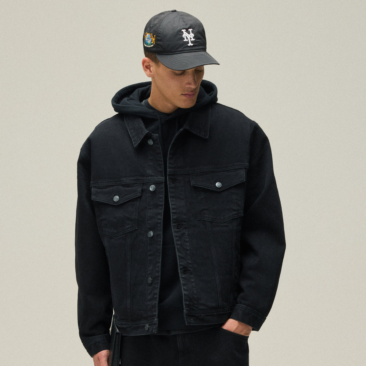Man in a black washed denim trucker jacket over a hoodie with a gray New Era NY cap, Kith Origins
