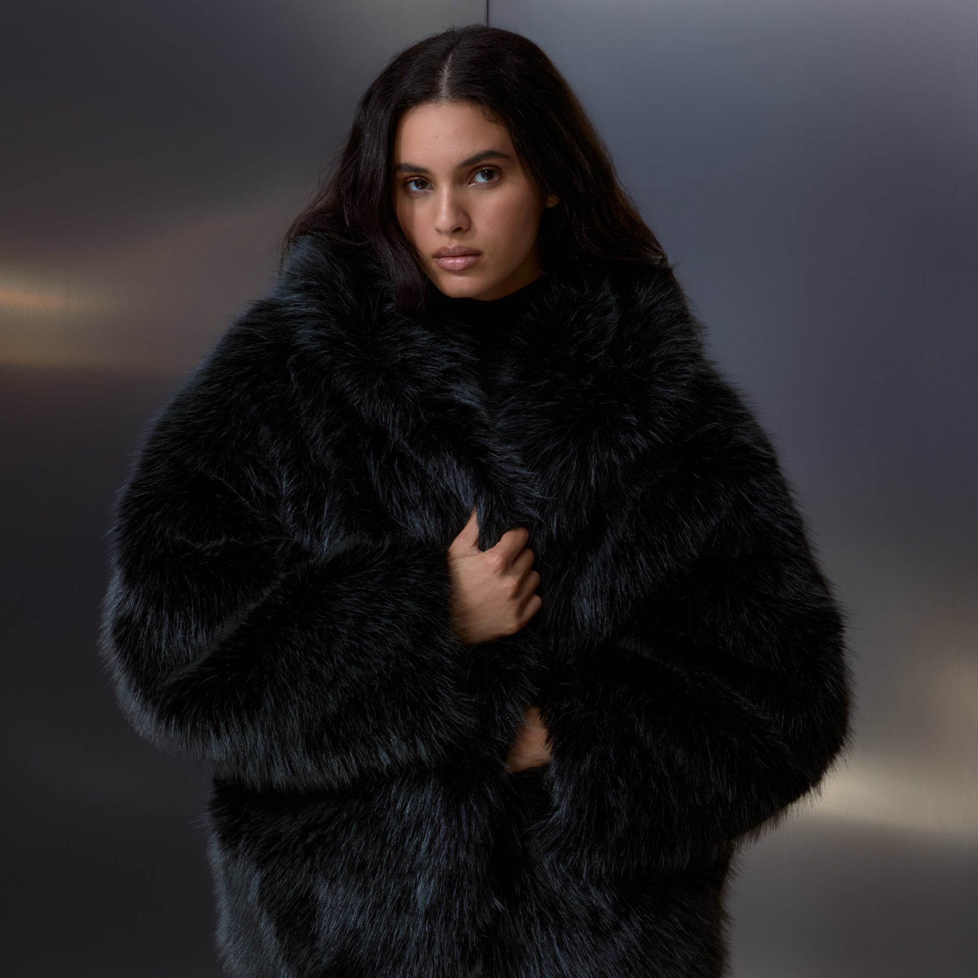 Woman wrapped in an oversized black shaggy faux-fur Kith coat over a black turtleneck against a moody gray studio backdrop