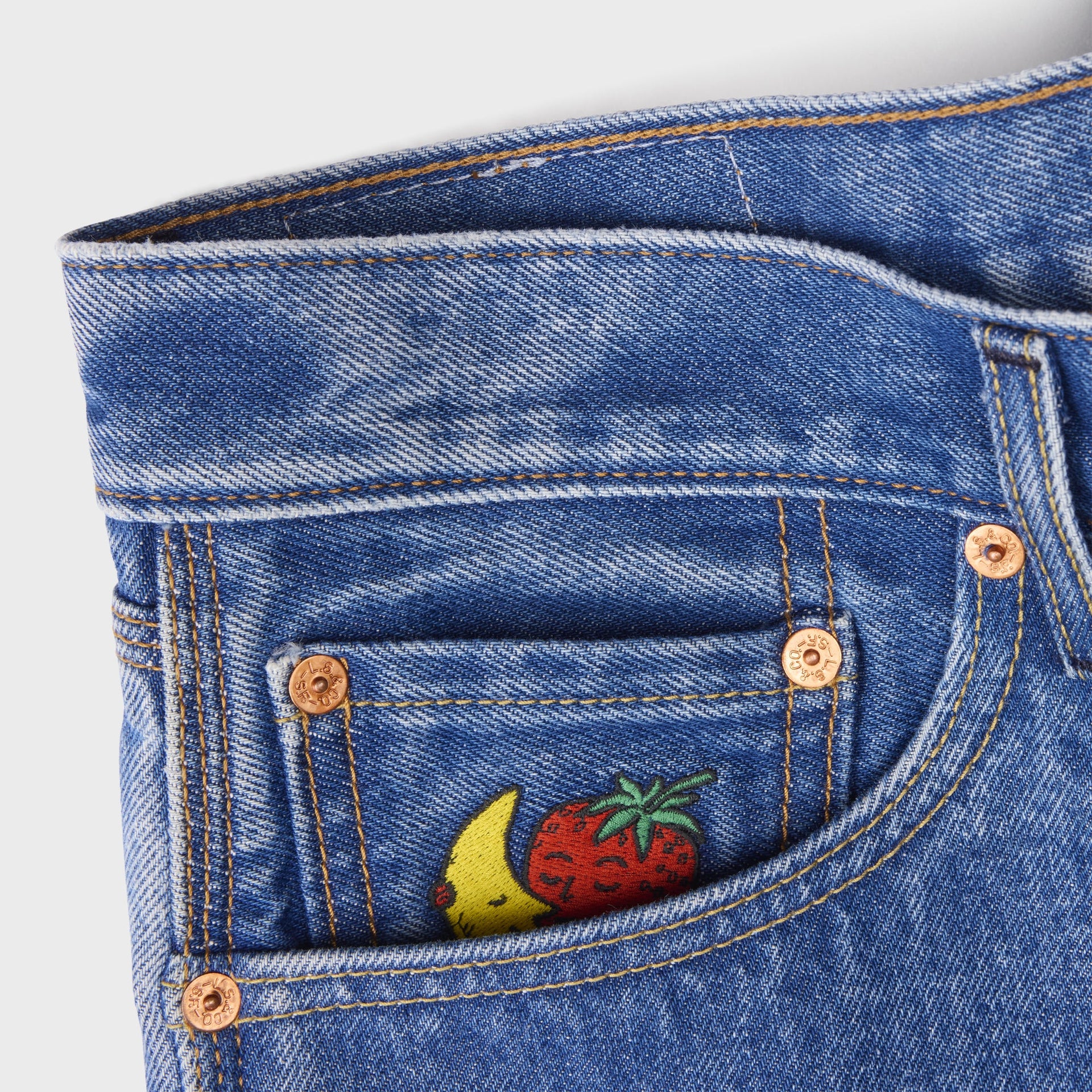 Detail of Levi's x Sky High Farm mid-indigo shorts front pocket with copper rivets and embroidered banana-and-strawberry mascot