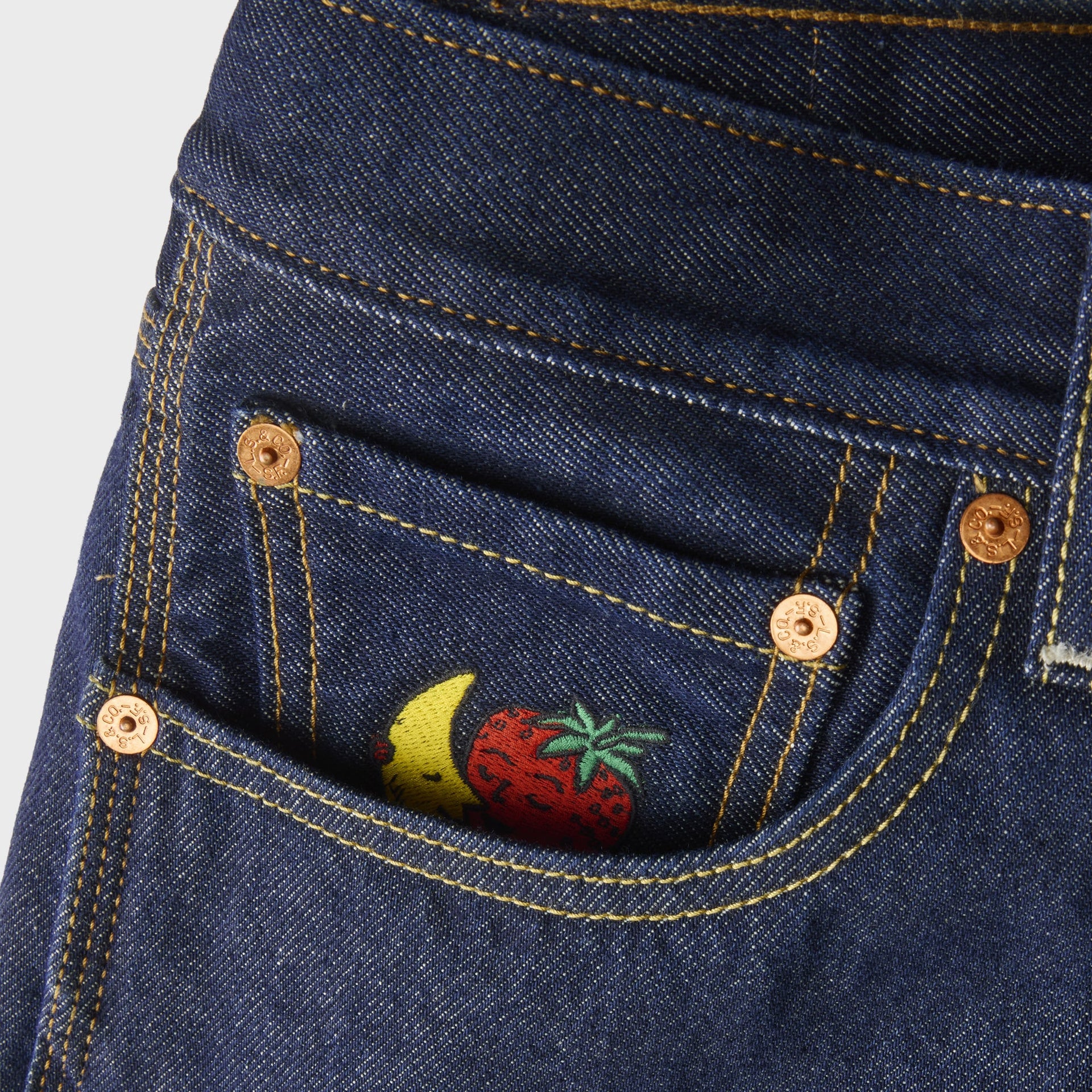 Detail of Levi's x Sky High Farm mid-indigo jeans front pocket with copper rivets and embroidered banana-and-strawberry mascot