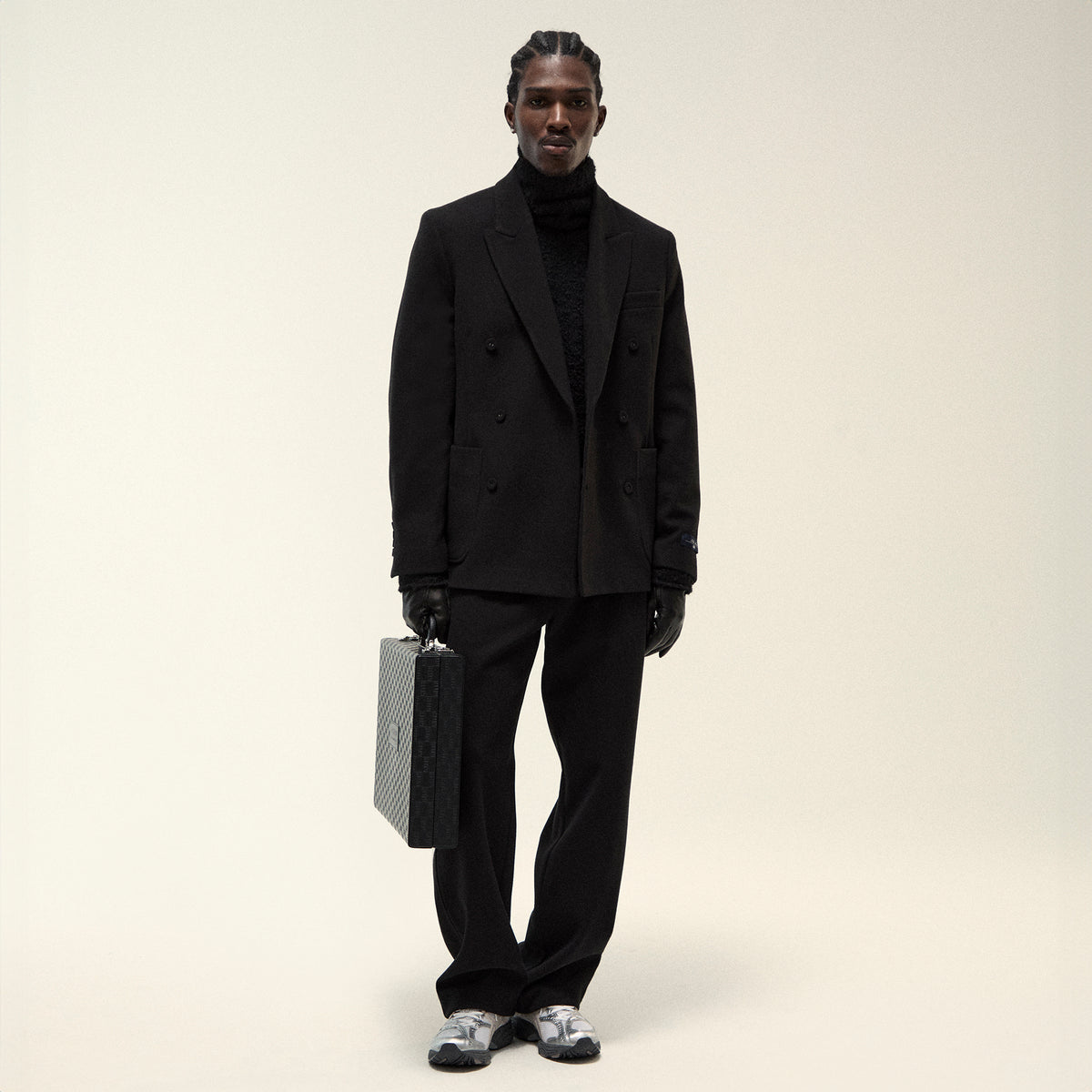 Model in black double-breasted wool blazer and turtleneck with wide-leg trousers, leather gloves, holding a monogram briefcase, silver sneakers