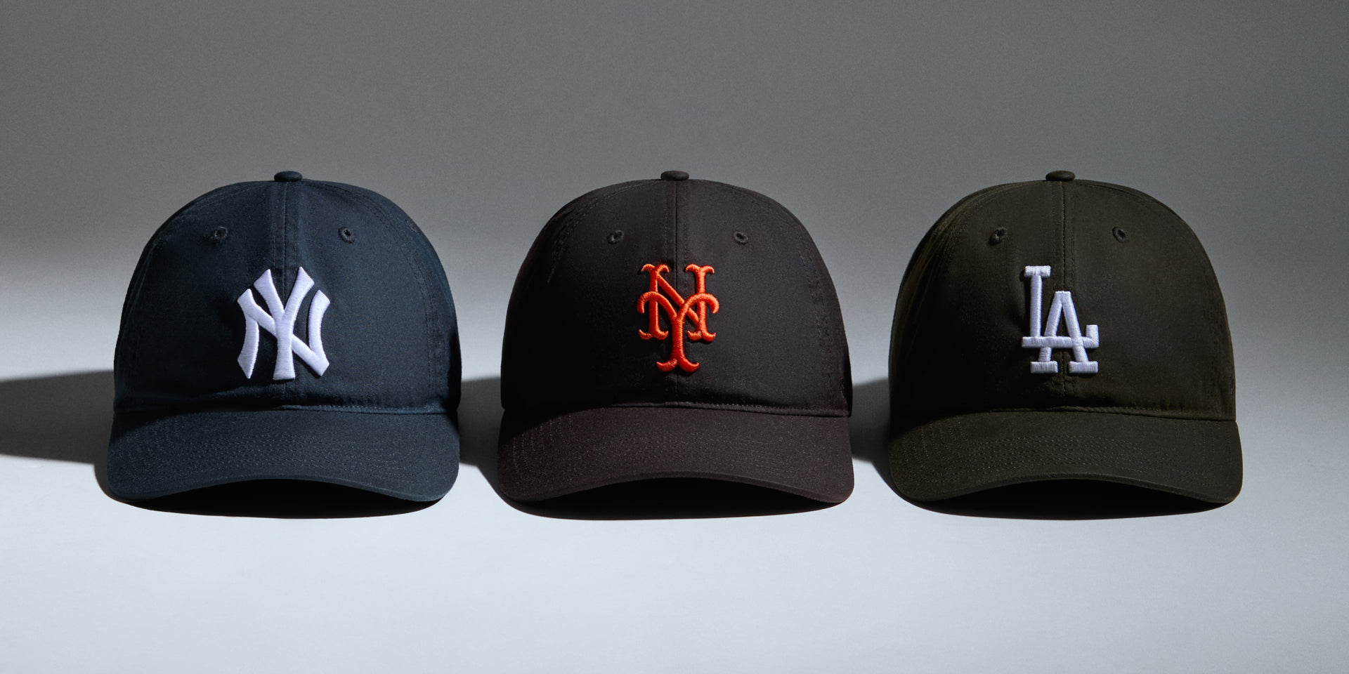 Three New Era ballcaps in a row: navy NY Yankees, black NY Mets and olive LA Dodgers on a grey studio backdrop.