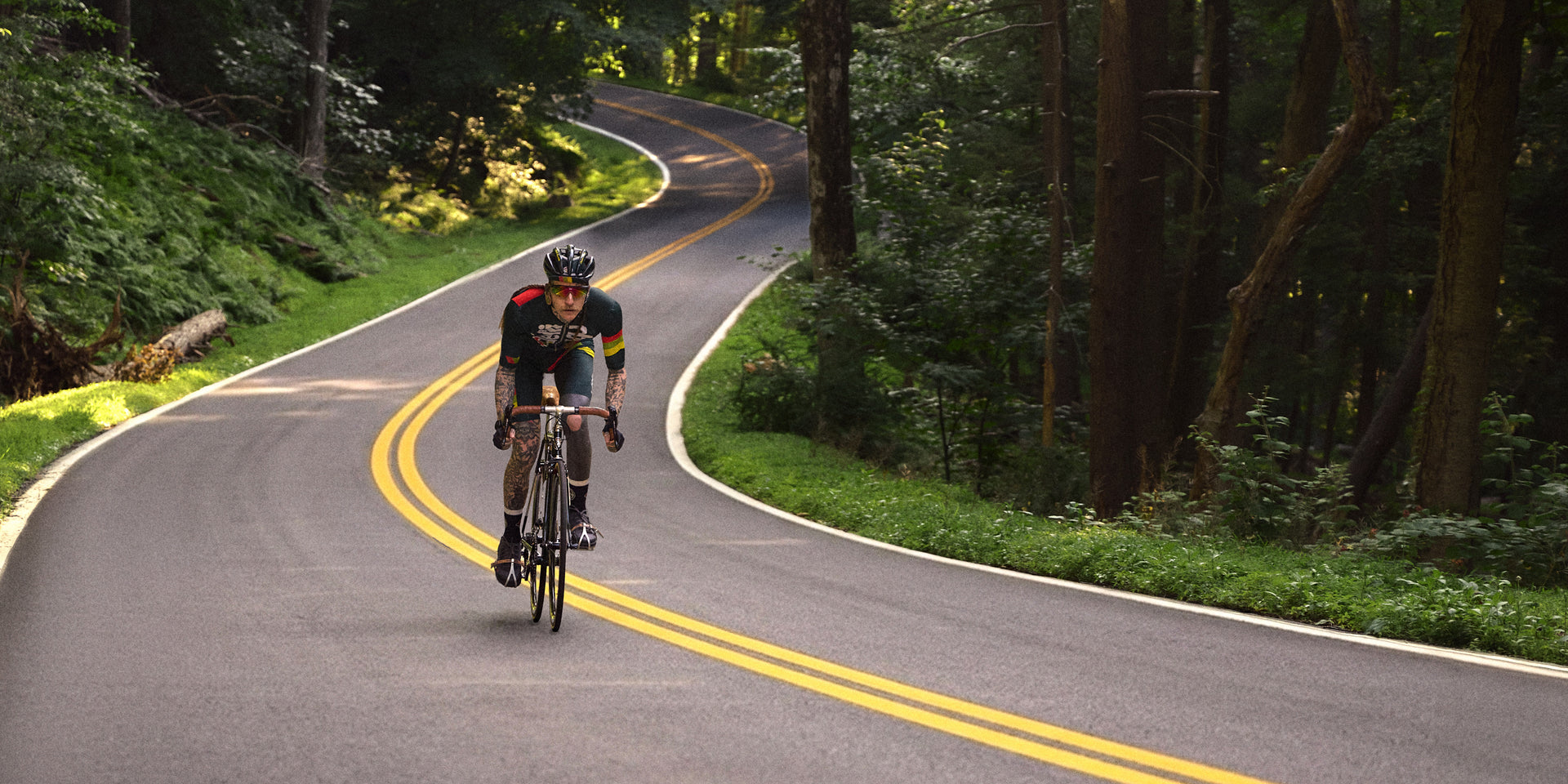 Tattooed cyclist in a dark green Kith Cycling jersey with red and yellow stripes riding a vintage road bike down a winding forest road with double yellow lines, wide crop