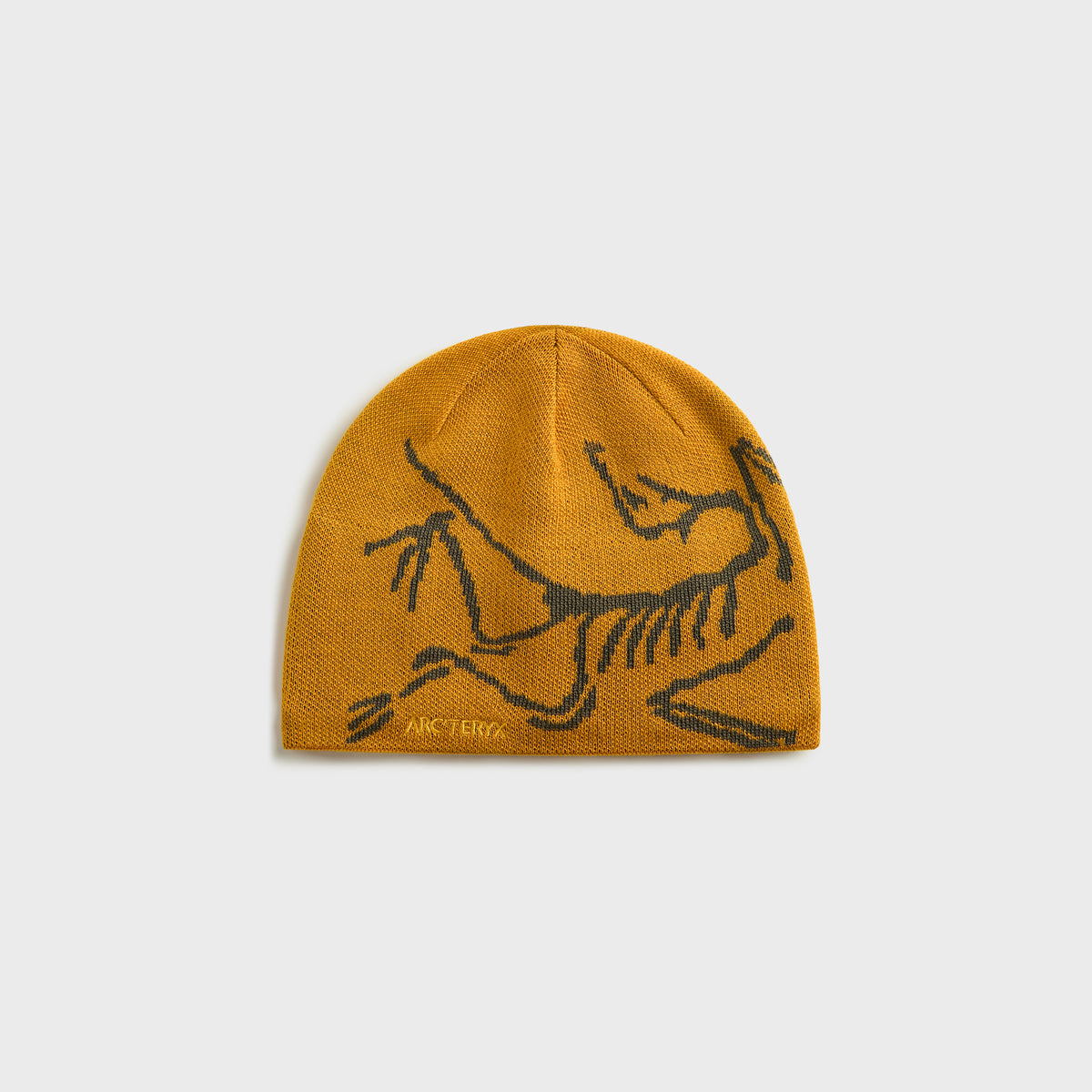 Arc'teryx mustard-yellow knit beanie with large grey Archaeopteryx bird logo graphic, front view on grey background