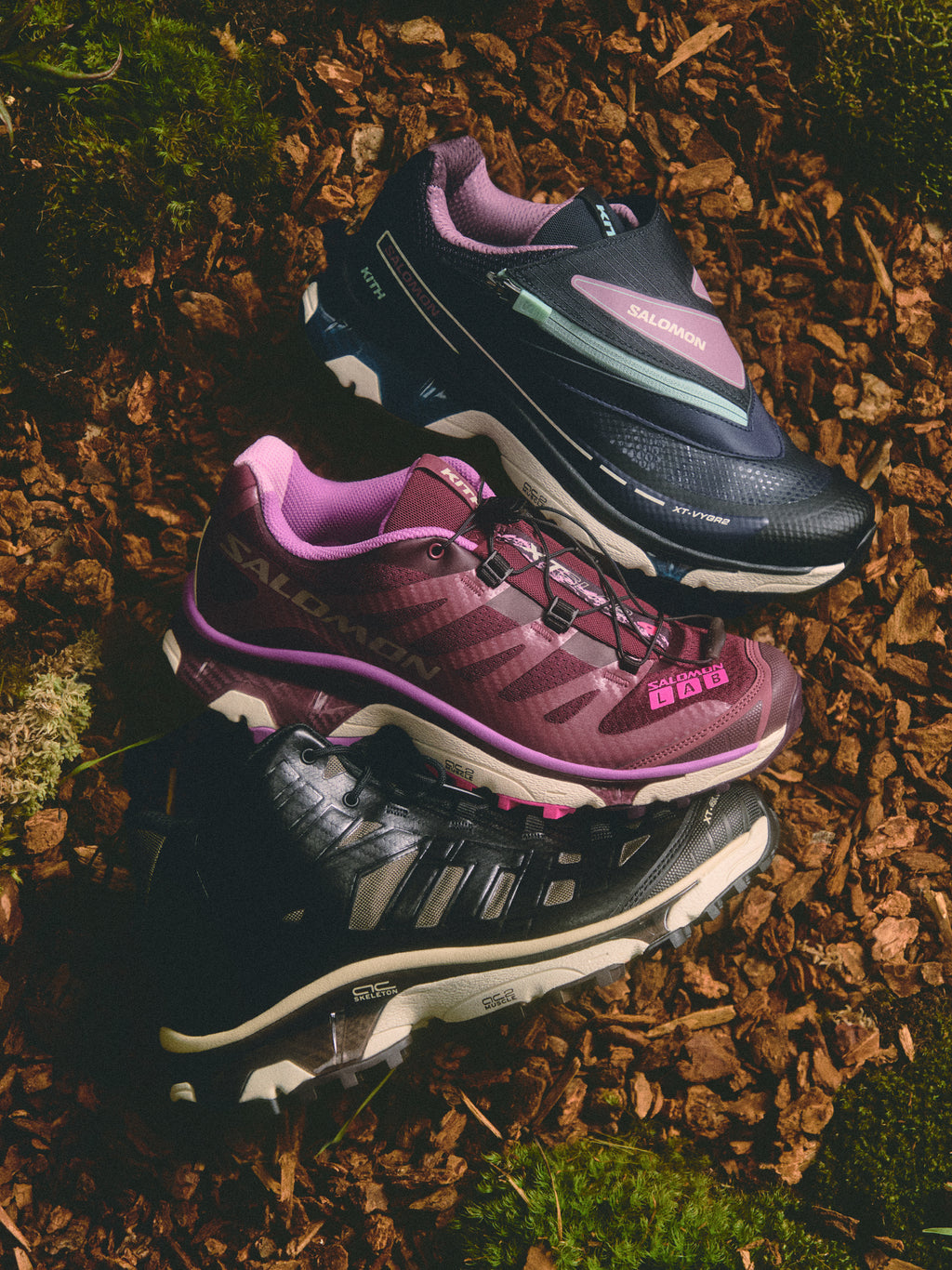 Three trail-style sneakers in navy, plum-pink and black laid on wood chips and moss, top-down editorial shot