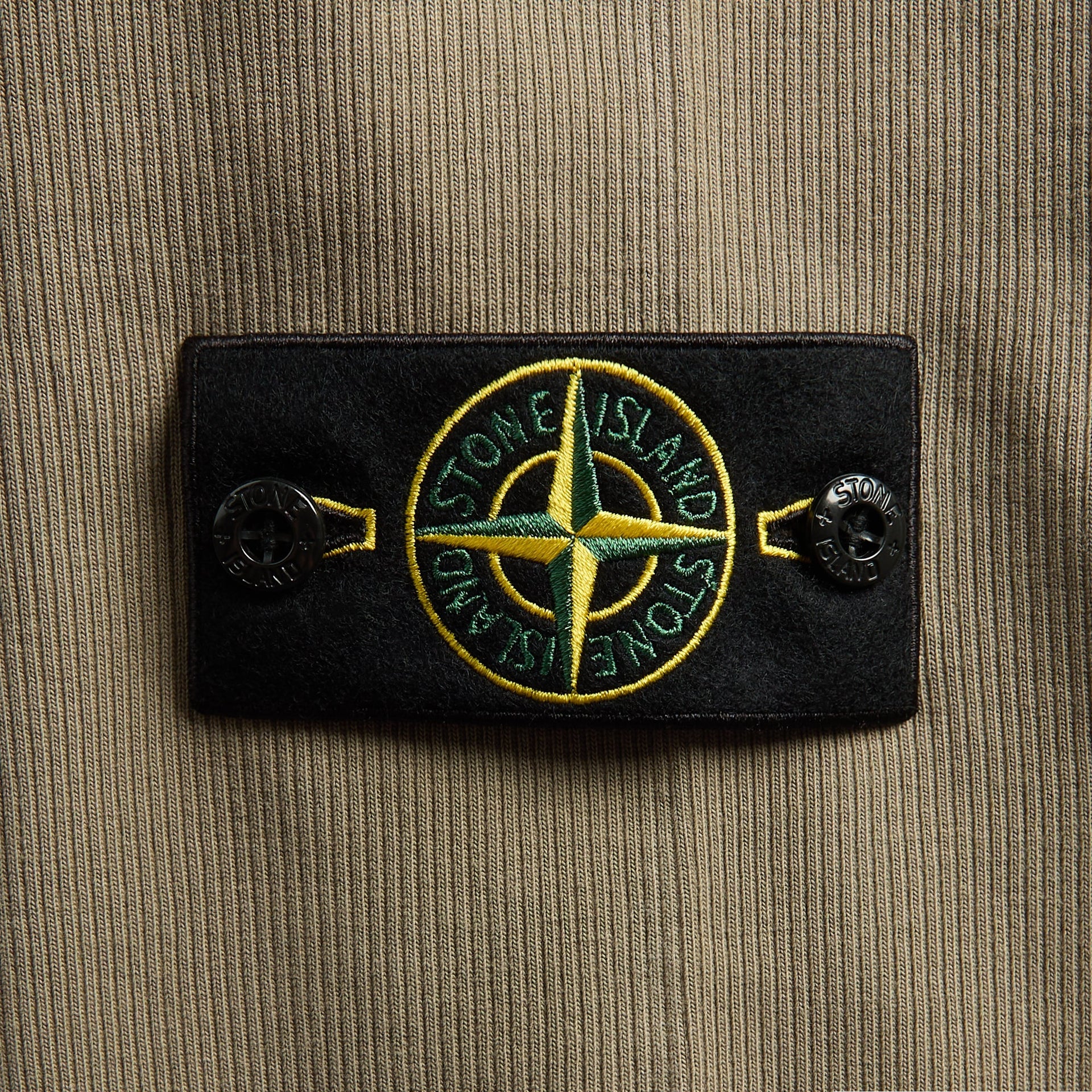 Close-up of Stone Island compass badge, black patch on military green ribbed sweatshirt sleeve