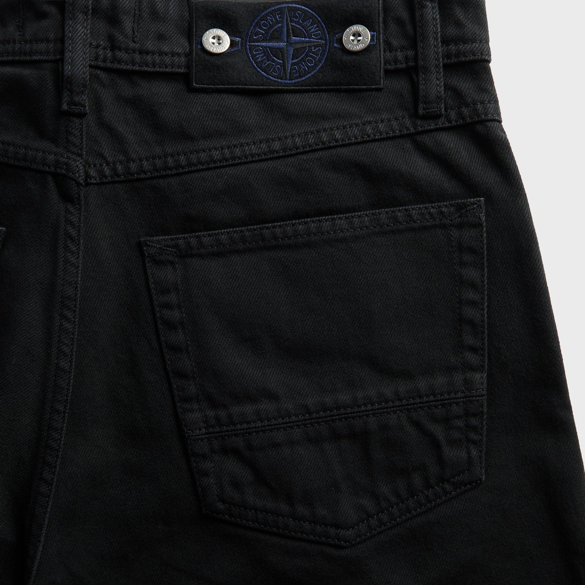 Detail of Stone Island navy-embroidered leather compass tab on black denim back pocket