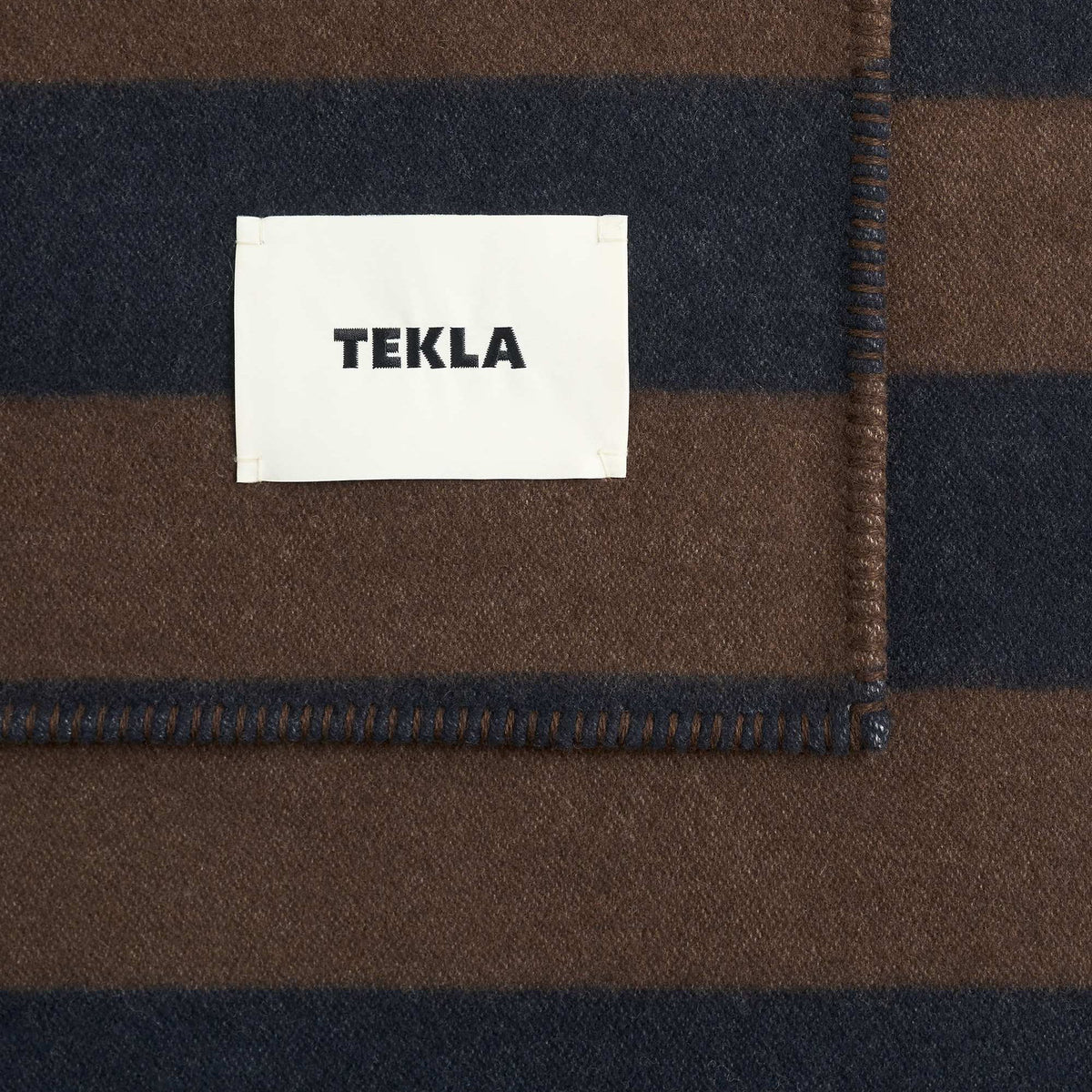 Tekla Lambswool Blanket in Walnut / Dusk Blue Stripes, close-up of corner with woven TEKLA label and stitched edge