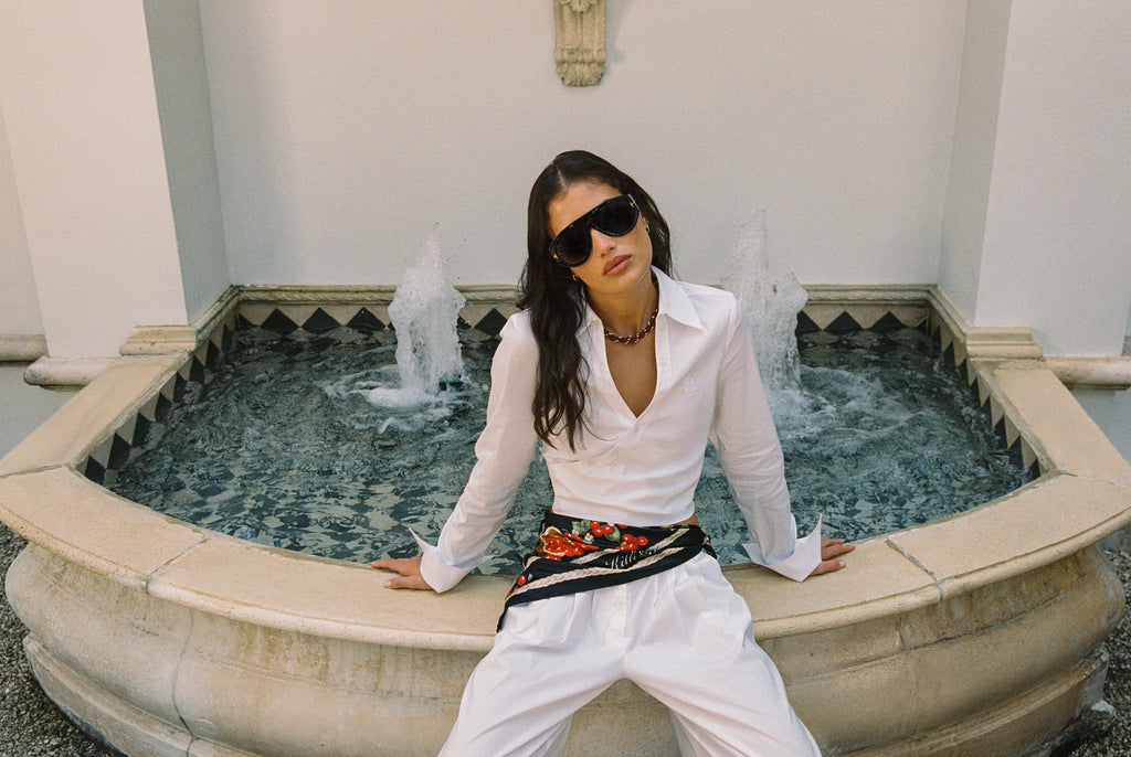 Woman in white Kith shirt and trousers with printed scarf belt seated at a stone fountain