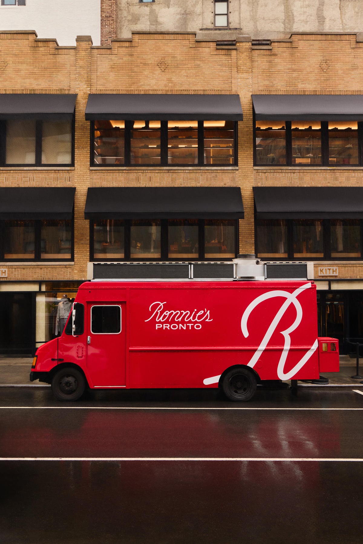 Ronnie's Pronto red food truck parked on a wet street below the Kith NYC storefront's brick facade and black awnings